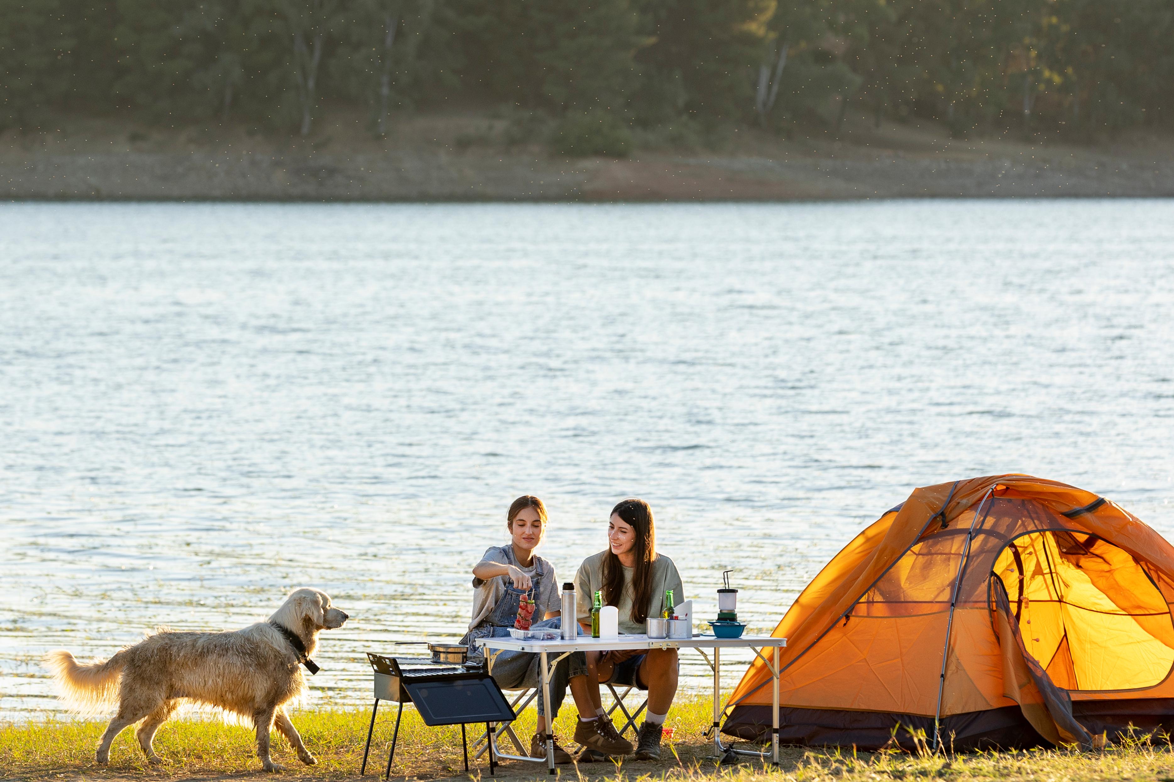 Lakeside camping scene with a tent, outdoor table, two campers and a dog